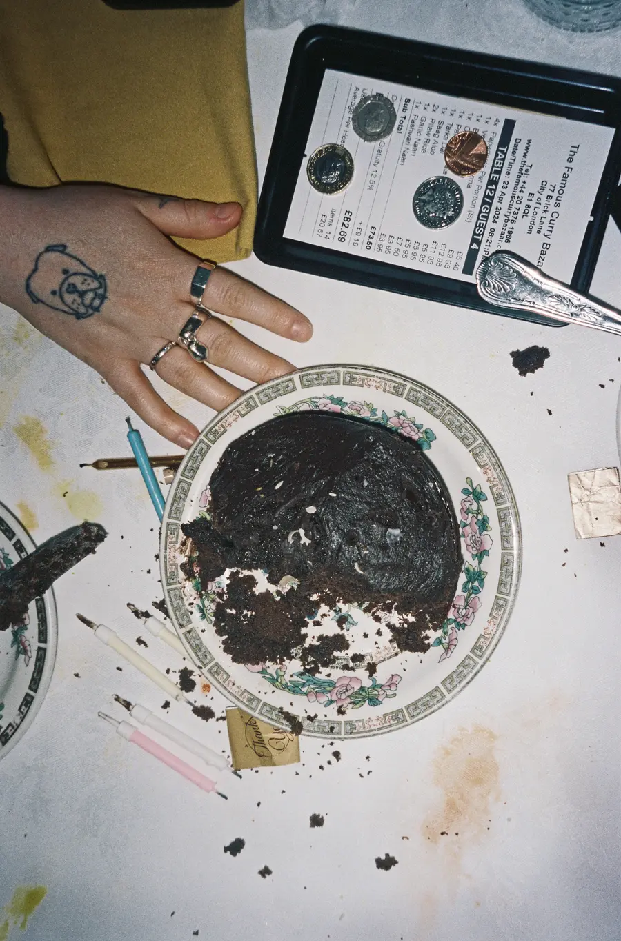  Colour photograph of a half-eaten  chocolate cake on a table. Alongside it, there is a menu,  some candles and a hand on the table as well.
