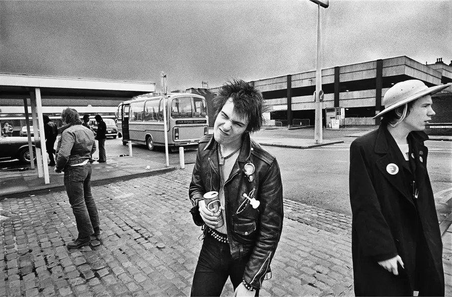 Black and white image of Sid Vicious and Johnny Rotten stood in a bus station casually strolling about. Sid holds a tin of beer in one hand and bares his teeth at the camera.