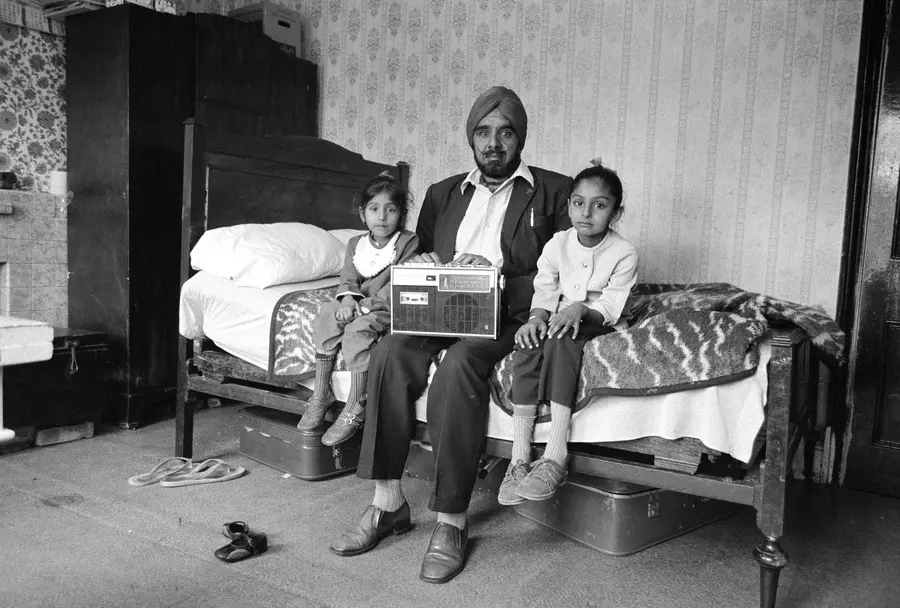Black and white photograph of a man sitting on a bed with his two small daughters sat on either side of him. A radio set is perched on the man’s lap.