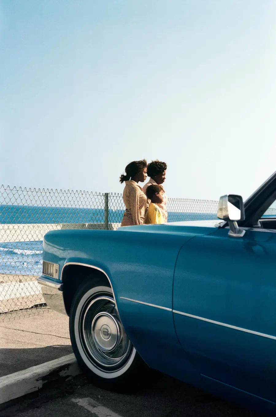 Colour photograph of two women and a young boy stood in front of a blue cadillac.