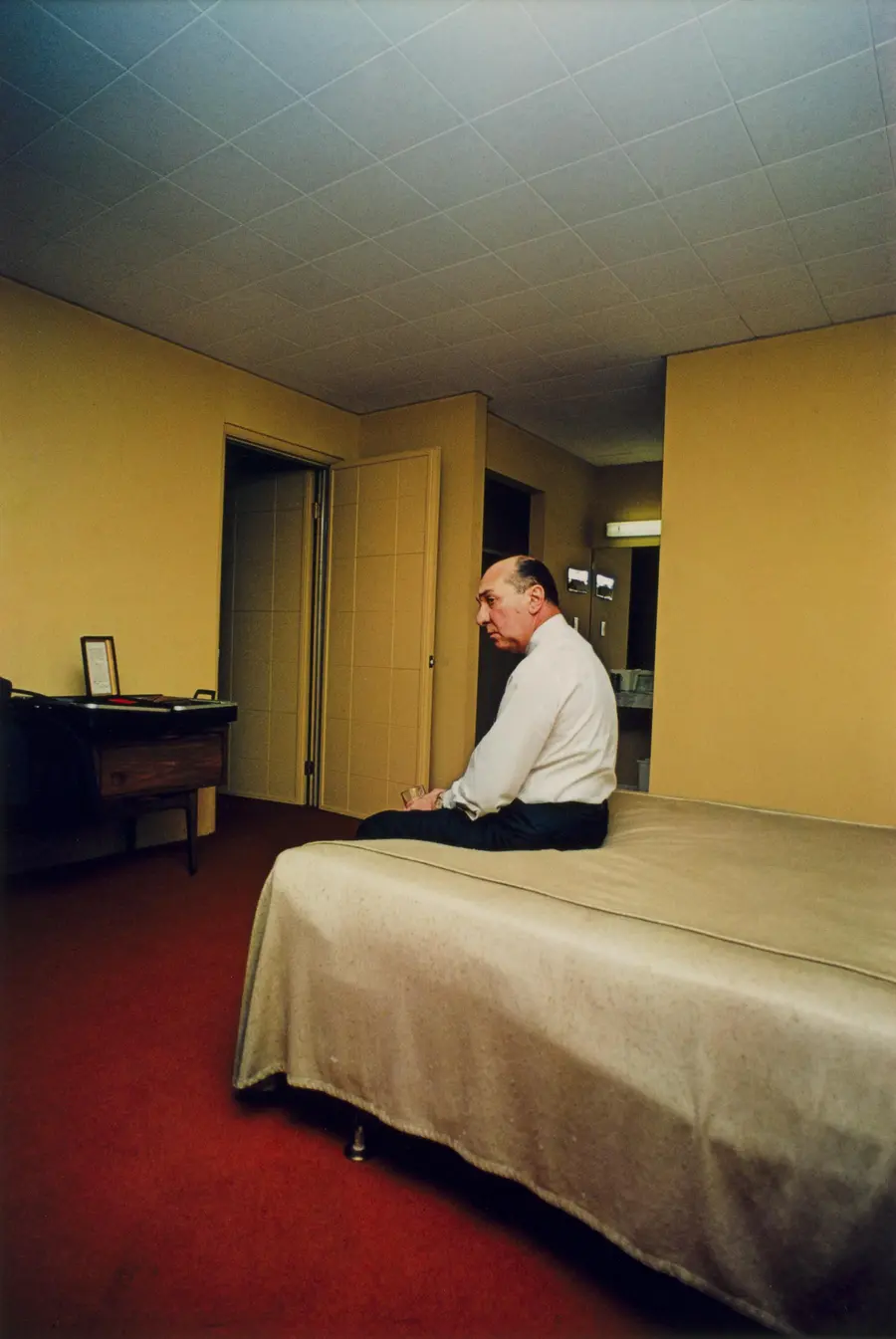 Colour photograph of a man sitting alone on the end of a bed in a room.