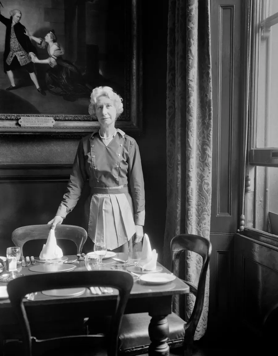 Black & white photograph of an older woman wearing a white apron standing by a window holding the back of a dining chair at set table.