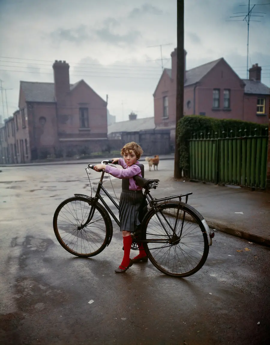 Colour photograph of a small child, straddling the frame of an adult-sized bicycle, who has stopped on a street and turned to look at the photographer.