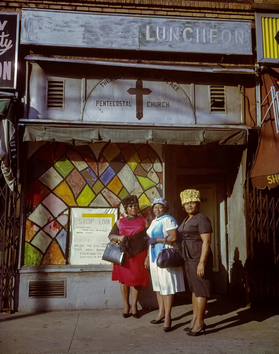A colour photograph of three women in their Sunday best standing outside of a Pentecostal church. 