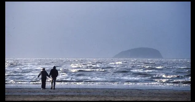 A photograph of a beach with 2 figures walking along the shore.
