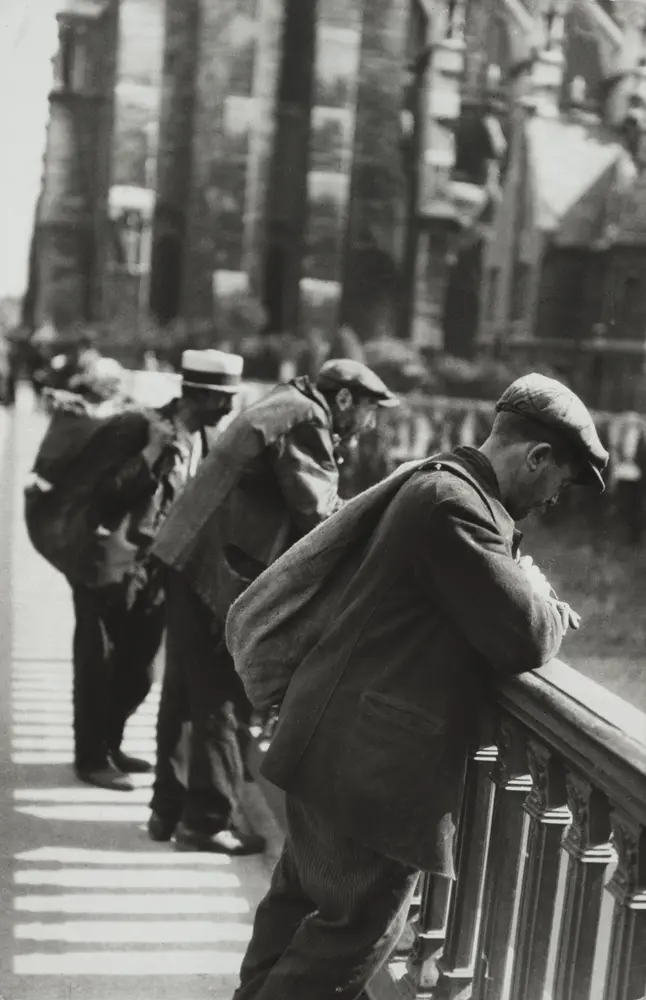 Three men lean over a bridge railing looking downward. 
