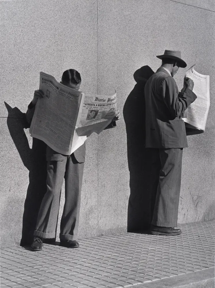 Black and white photograph of two men standing against a wall, each looking in a newspaper.