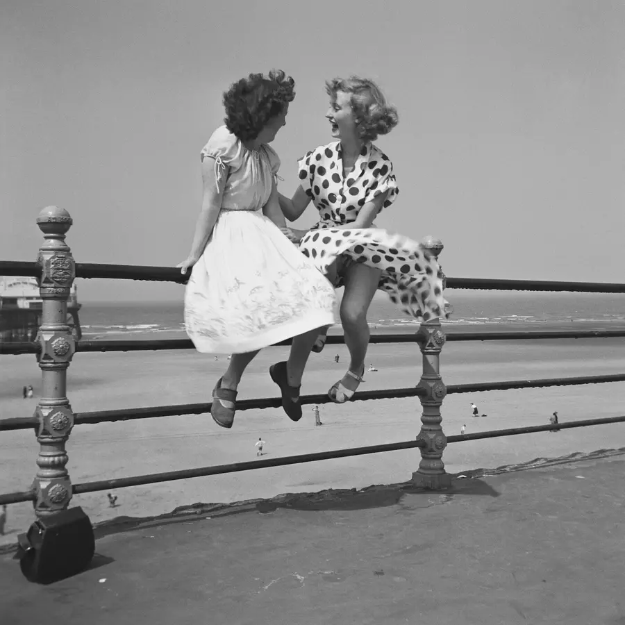 Black and white photo of two womenchatting on the railings by a beach.