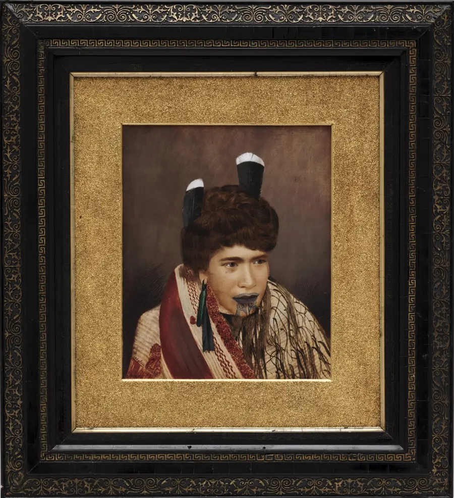 Gold and black framed, hand coloured black and white photograph of a woman in ceremonial dress with hand drawn markings on her lips and chin.
