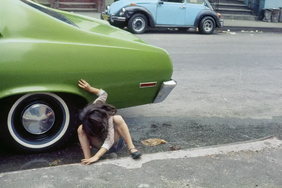 A child crouches on the pavement next to the back tyre of a green car