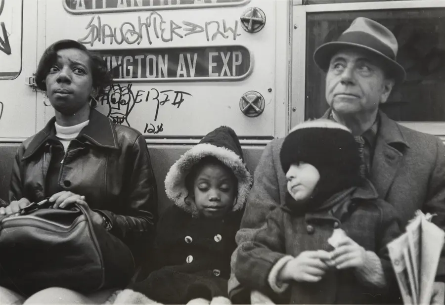 Two adults, two children are seated side-by-side on the subway, all facing the camera