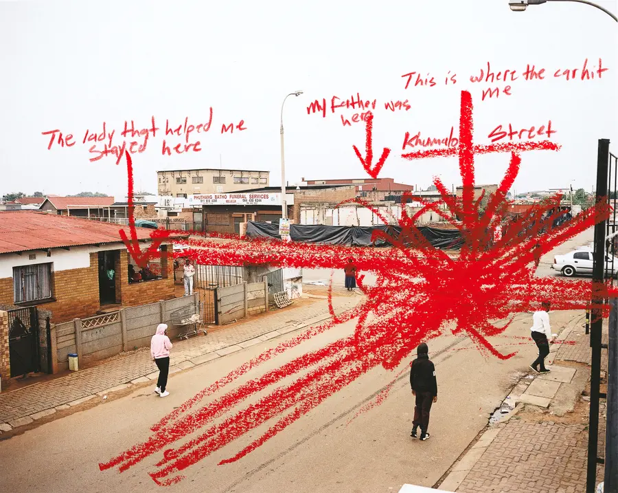 Colour photograph of people stood on a street. The photo is marked with red annotations saying: “The lady that helped me stay here”, “My father was here”, “This is where the car hit me” and “Khumalo Street.”