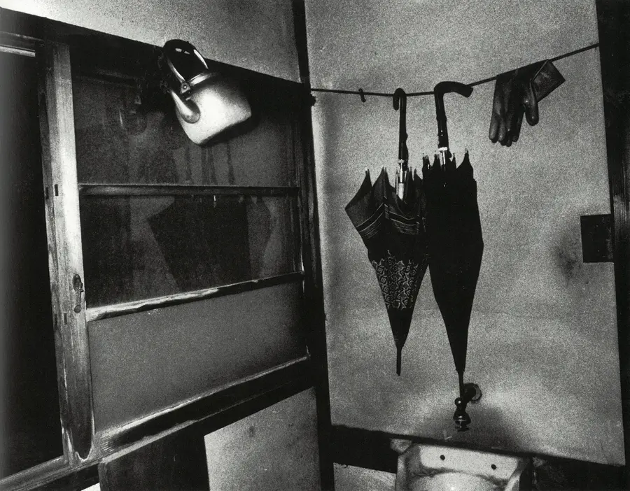 A black and white vintage photograph of a small room. A washing line hangs across it. A metal teapot and two umbrellas hang from the line.