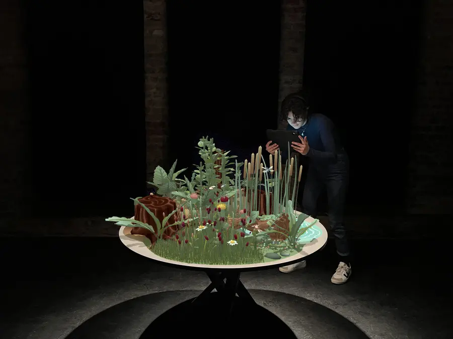 A young woman points a tablet at a cgi landscape of a natural ecosystem including daisies, leaves, logs, and water appears on a round table in a dark space