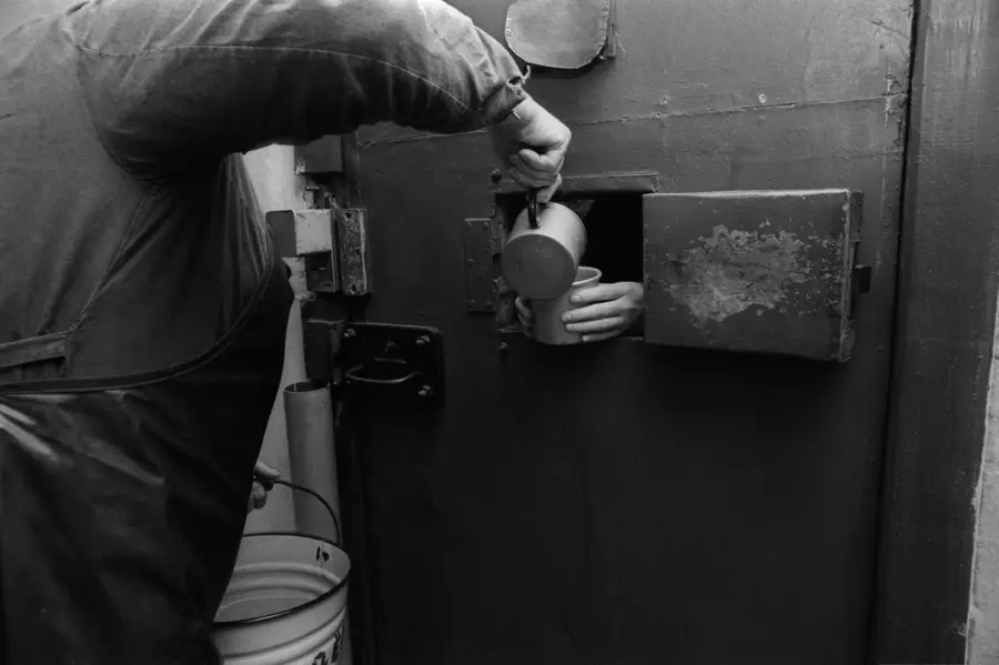 Black and white photograph of someone  pouring liquid into a cup held by a prisoner through a small  opening in a door