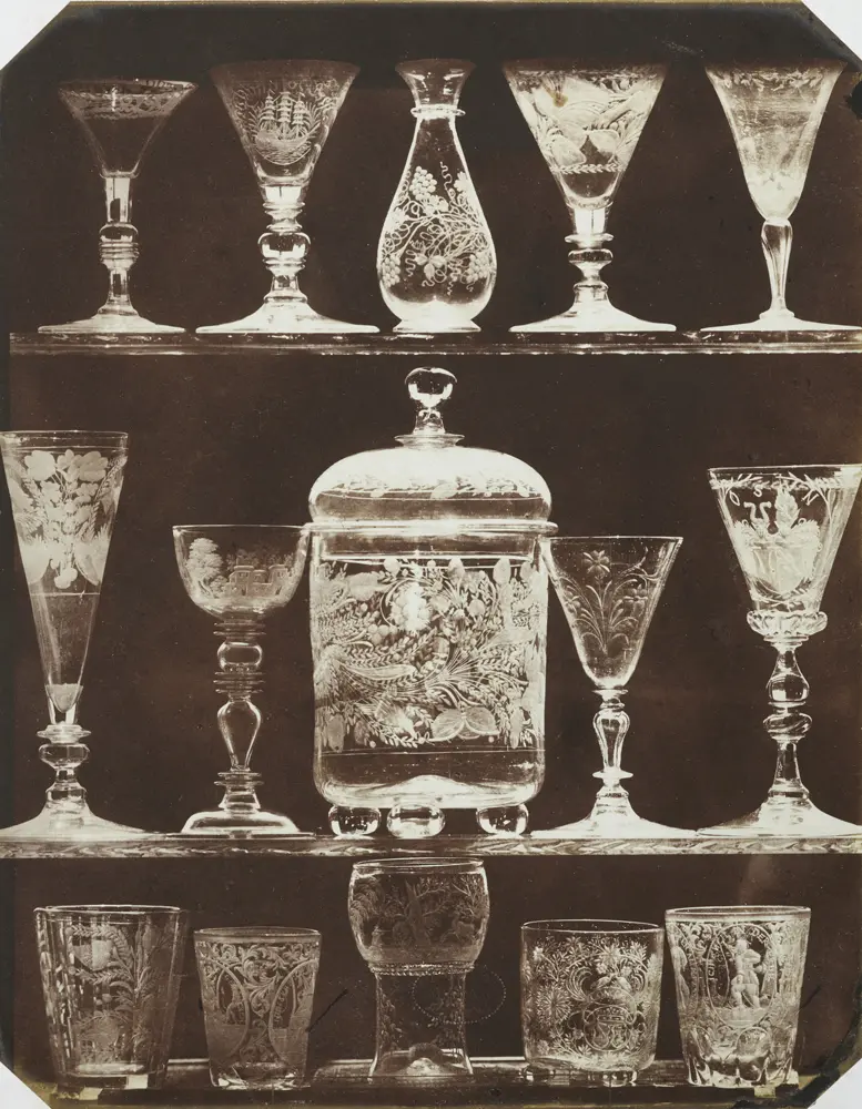 A brown monochrome photograph of sherry glasses and other glassware lined up on a shelf.