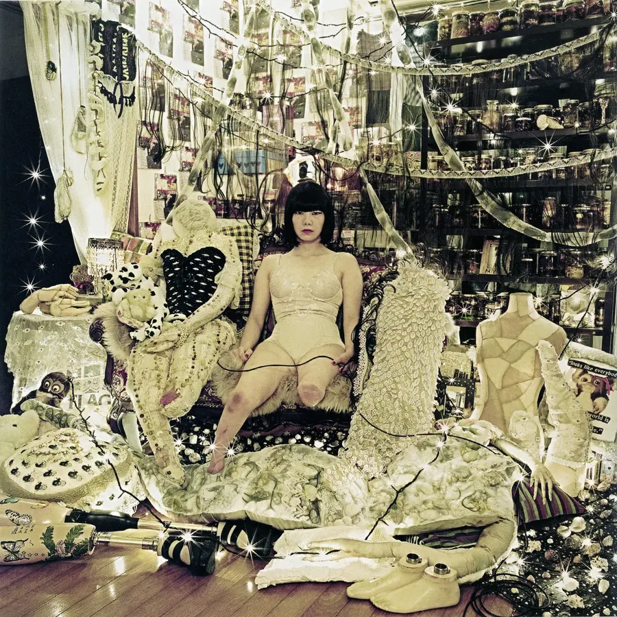 A woman sits in a workshop surrounded by thread organised on shelves and dressmaking dolls. She has both legs missing up to the knee. She looks directly into the camera with an assertive gaze.