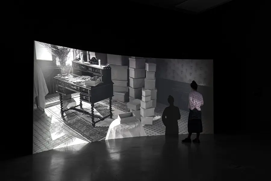 Installation photo of a screen showing a room full of boxes. A person can be seen watching this screen.
