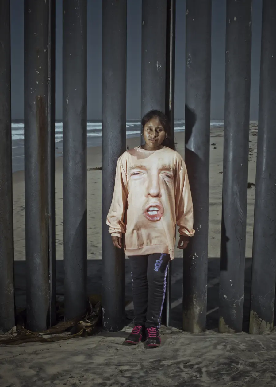 Colour photograph of a girl stood on a beach against a railing, she wears a t-shirt with Donald Trump’s face on it.