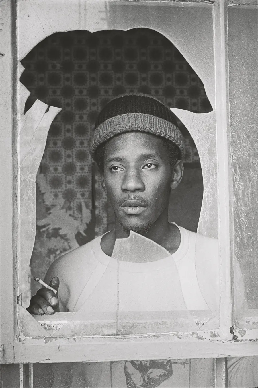 A black and white image of a young Linton Kwesi Johnson gazing out of a broken window pane.