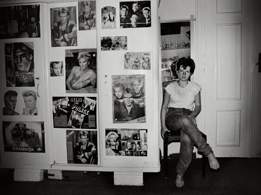 Black and white photograph of a young  woman sitting on a chair next to a wardrobe decorated  with several posters of musician Sting, and rock band ‘The Police.’