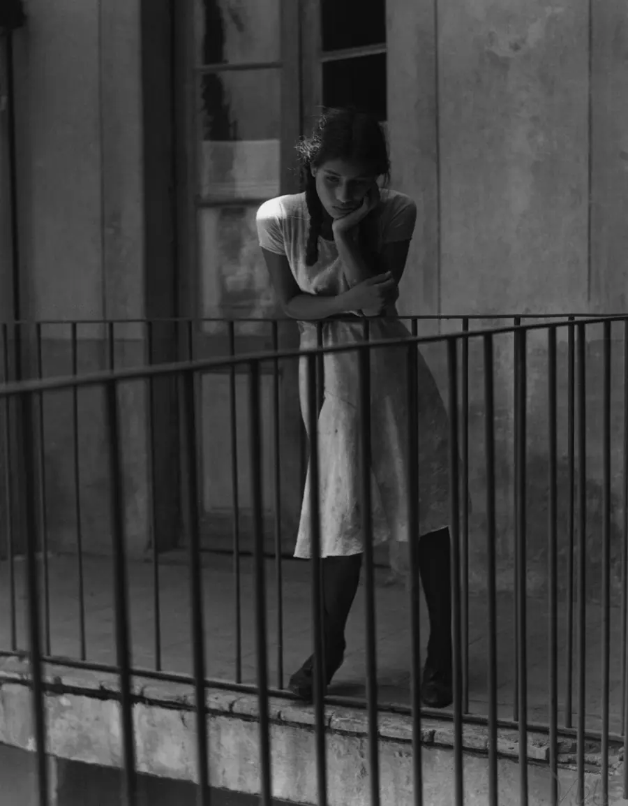 A black and white image of young girl with a contemplative expression  in a white dress looking over the railings of a staircase.
