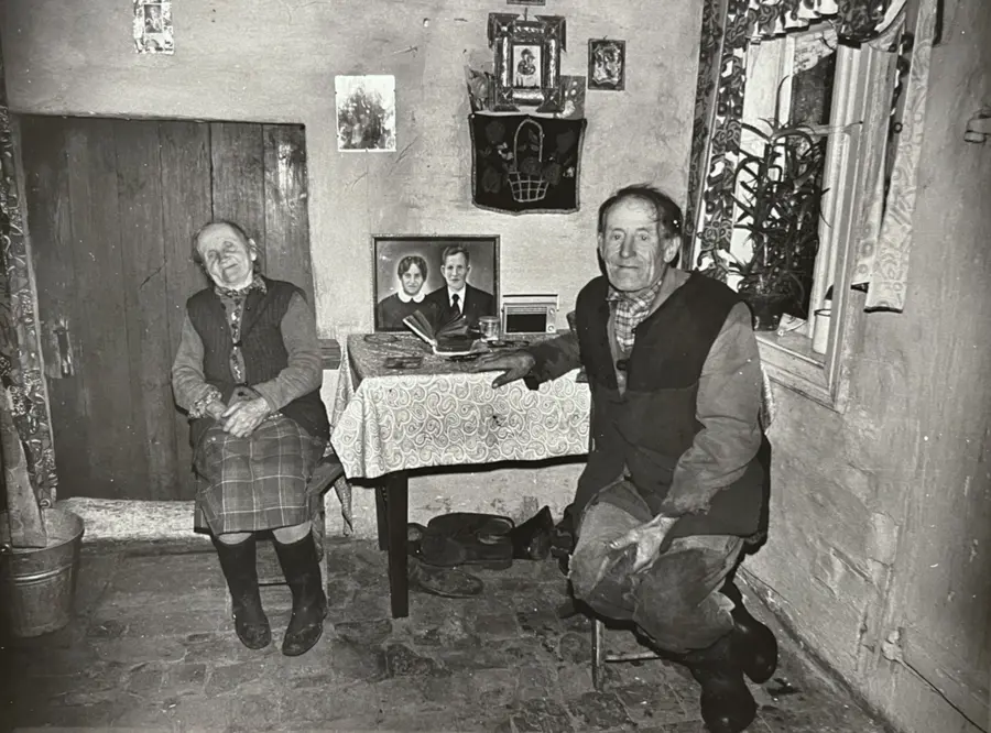 Black and white photograph of an elderly Polish couple sat at a dining table both with their chair facing the camera. They are in a small dining room and a photograph of their wedding day is propped up on the table sat in between them. Their portrait shows them in their youth dressed in their wedding clothes.