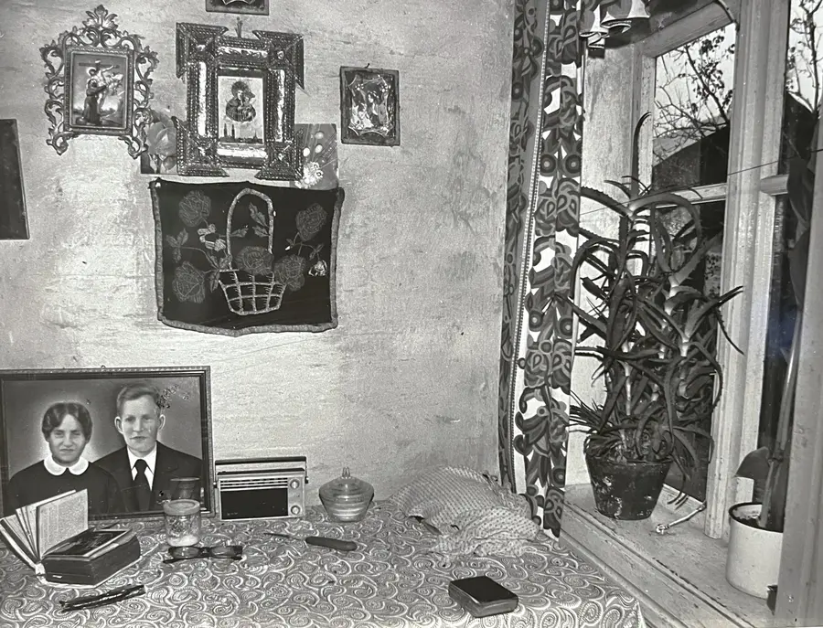 A black and white vintage photograph of an empty kitchen in rural Poland. A small window overlooking trees is to the right and the table covered in a paisley cloth occupies the lower half of the image. A portrait in a frame sits on the table featuring a man and woman posed in formal wear on their wedding day.
