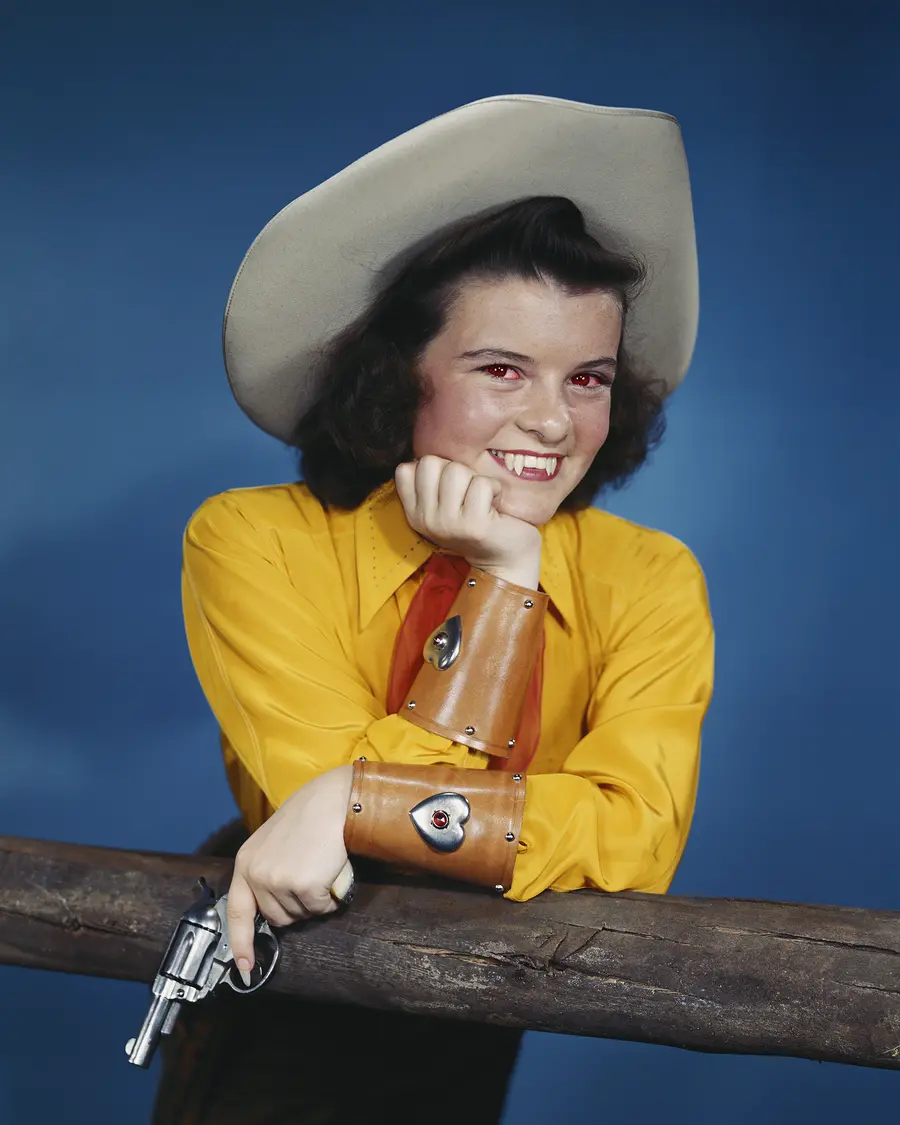 Colour photograph of a woman in cowboy attire posing with a gun in her hand. In the picture you can also see she has extended incisors and reddened eyes.