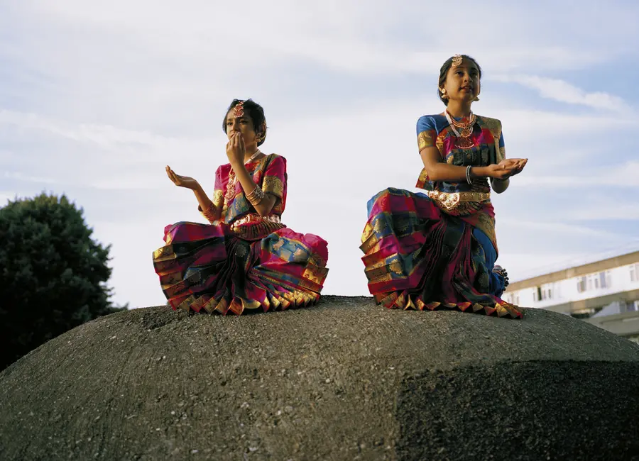 A pair of young desi women dressed in traditional clothing are shot mid-dance in a crouching pose.