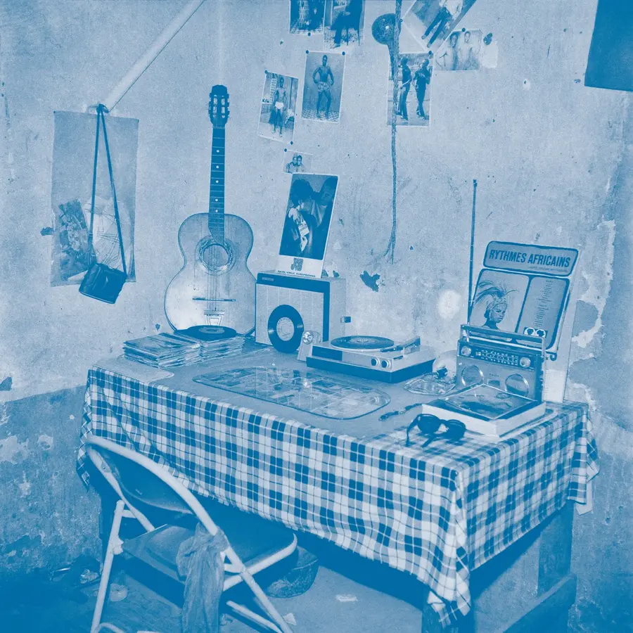 A vintage greyscale image of a desk with a record player, records and a guitar resting on top of it amongst other paraphernalia. 