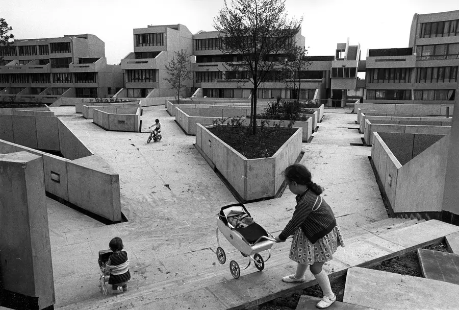 Black and white photograph of two children pushing toy prams in a concrete, open space with stairs, and another child riding a bicycle outside a housing complex. 