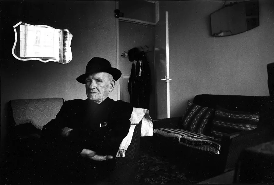 Black and white photograph of person sitting in a chair in a flat, arms folded looking to camera.