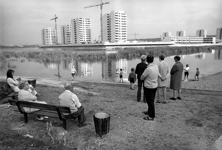 Black and white photograph of people by the bay of a pond surrounded by construction sites