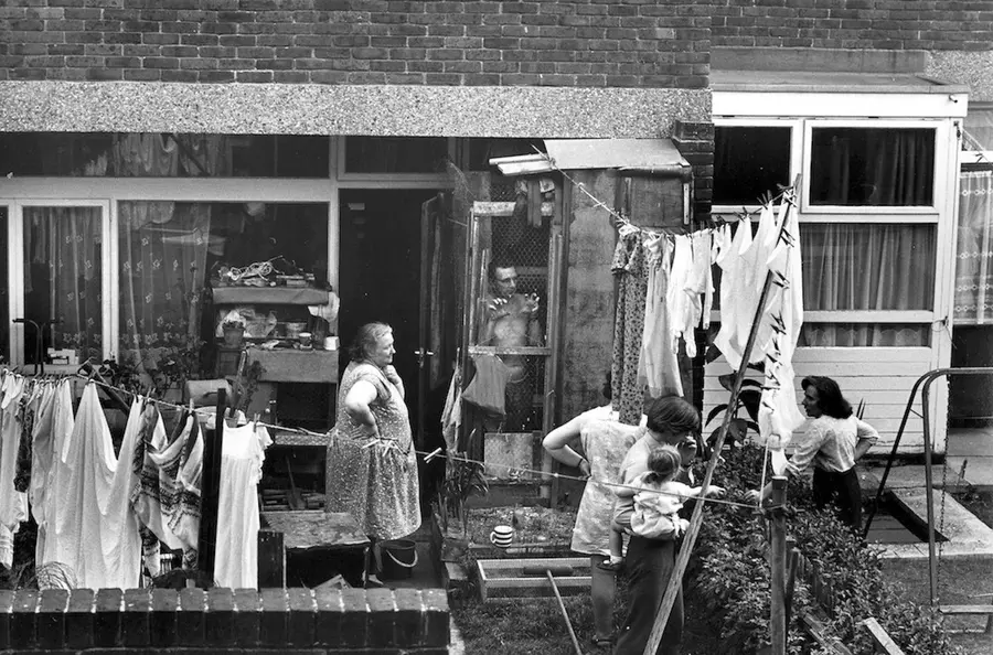 Black and white photograph of neighbours speaking to each other over a garden wall.
