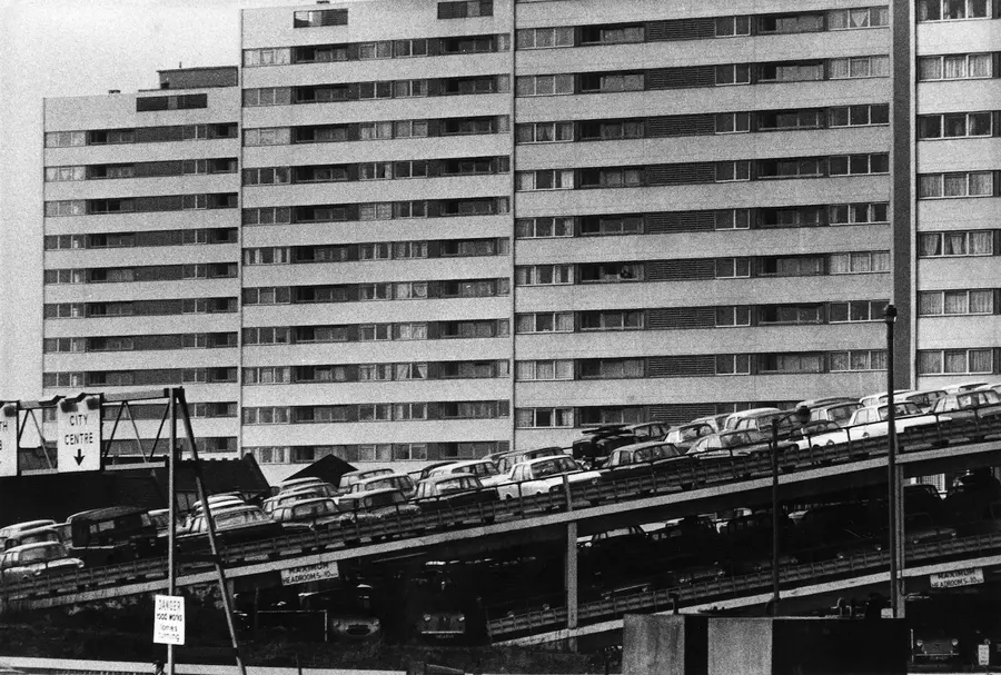 Black and white photograph of a multi-story parking lot and road, angled upward from left to right, in front of a tall block of flats nearly filling the background.