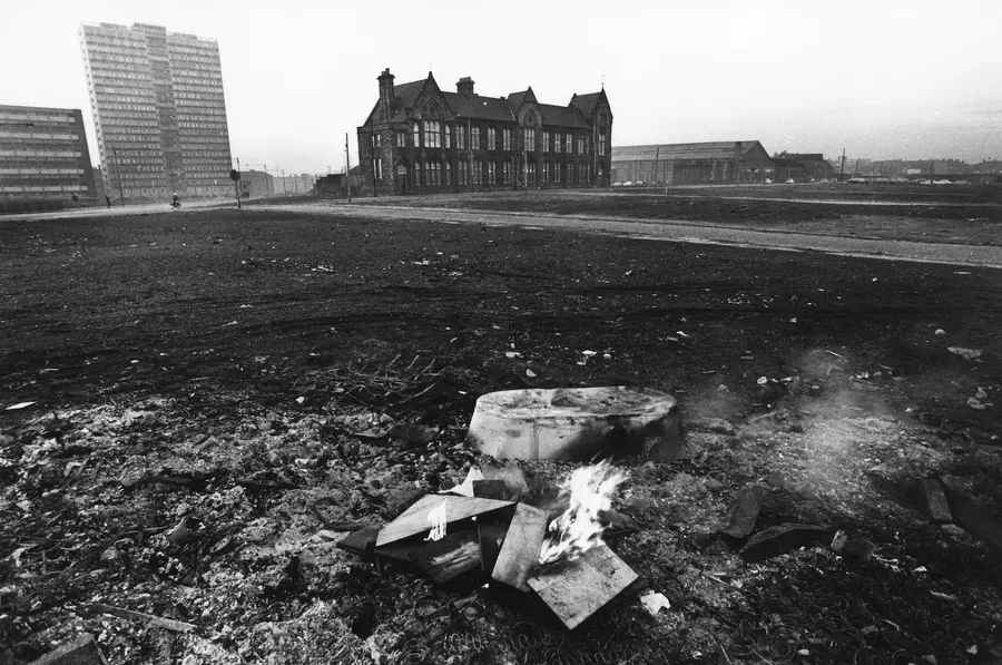 Black and white photograph of a landscape, with an upturned tin bath and fire in the foreground, flattened earth in the middle ground, and a Victorian school and high rises in the background.