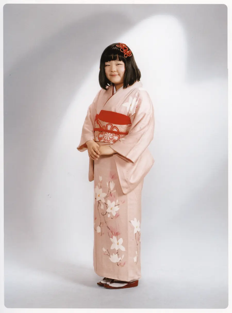 A woman in a pale pink kimono stands posing in a white photo studio.