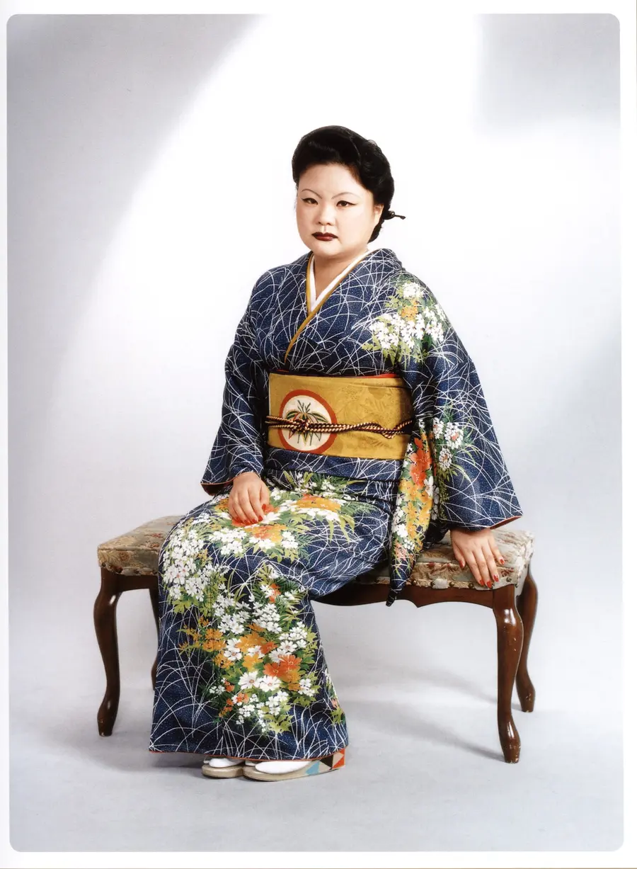 A woman in a dark blue patterned kimono is sat on an upholstered wooden stool in a white photo studio.