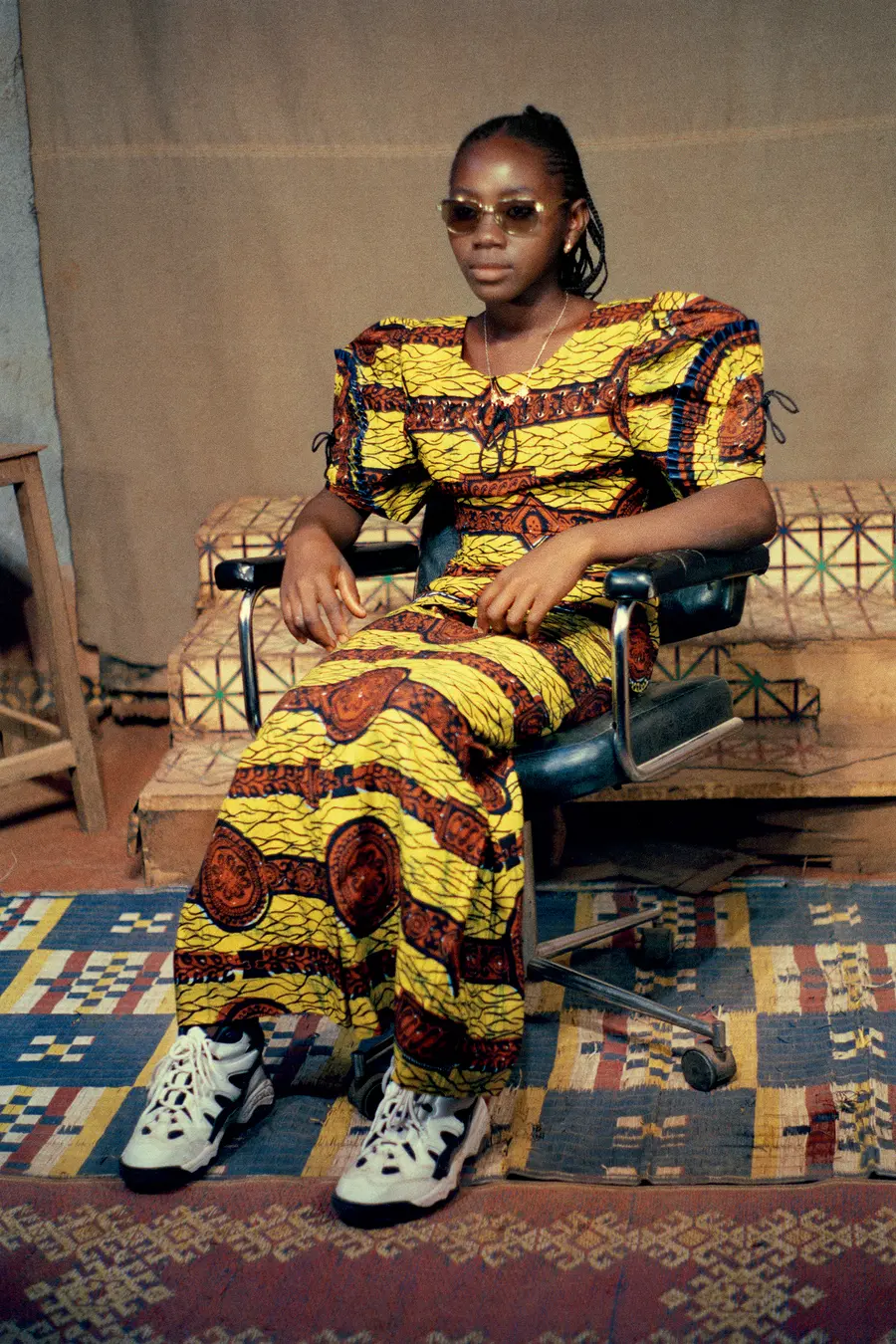 Colour photograph of a woman sat on a  chair. She wears a traditional dress with sunglasses, and  trainers.