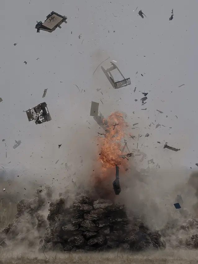 Colour film still of an explosion with debris flying in the air. In the centre a fire burns.