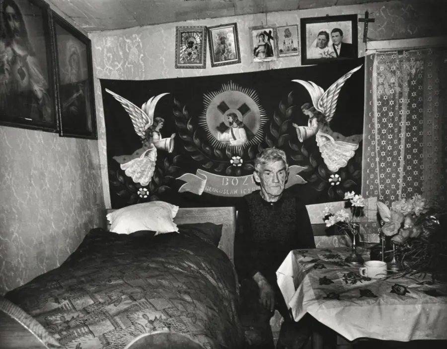 Black and white photograph of an older  man sat in the middle of a bedroom. Family photographs  and religious ephemera can be seen on the walls around him
