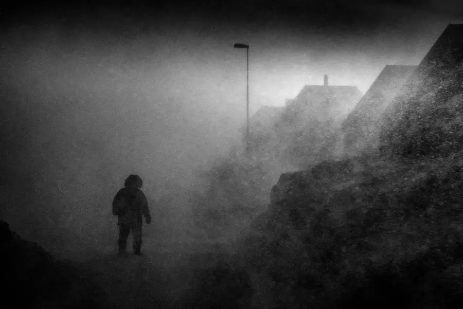 A greyscale scene of a heavy snow storm. I figure in a  thick winter coat walks through it under the light of a lamp post.