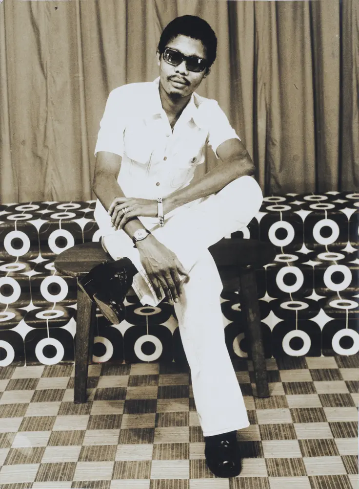 Photograph full length portrait of a seated man with a short afro and goatee, wearing dark sunglasses and seated in a boldly patterned studio.