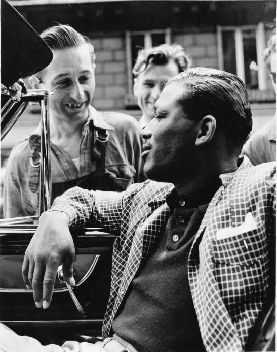 Black and white photograph of a person sat in a car with the window down and two people outside the car looking in and smiling