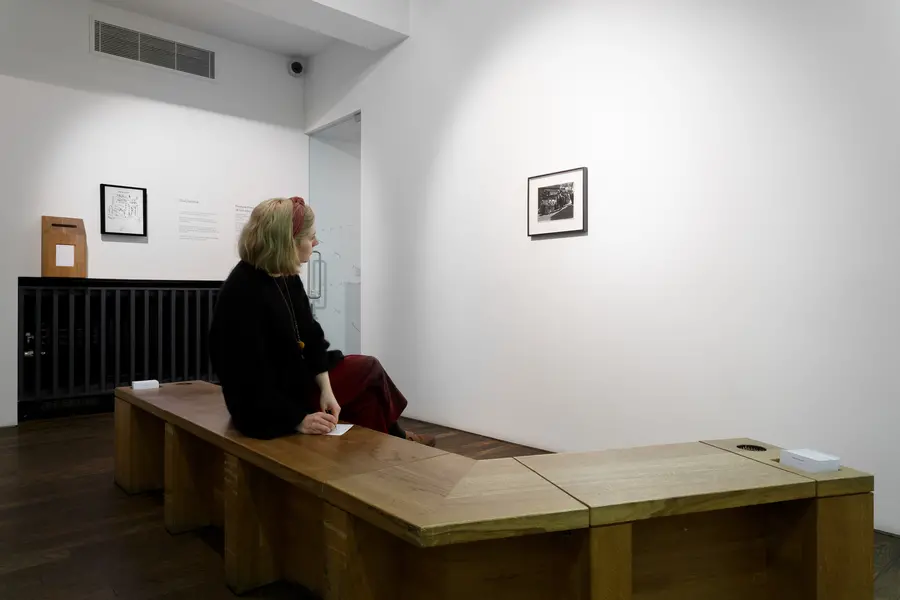 Person on a bench writing on a postcard while looking at framed black and white photograph on wall.
