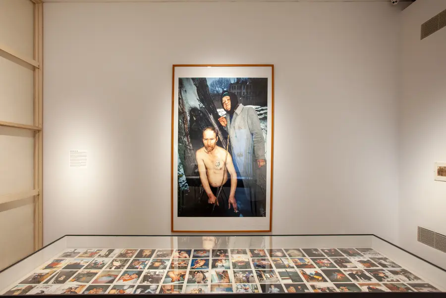 A view of an exhibition with one framed photo of a shirtless man stood next to a woman in a heavy overcoat in the snowy woods.
