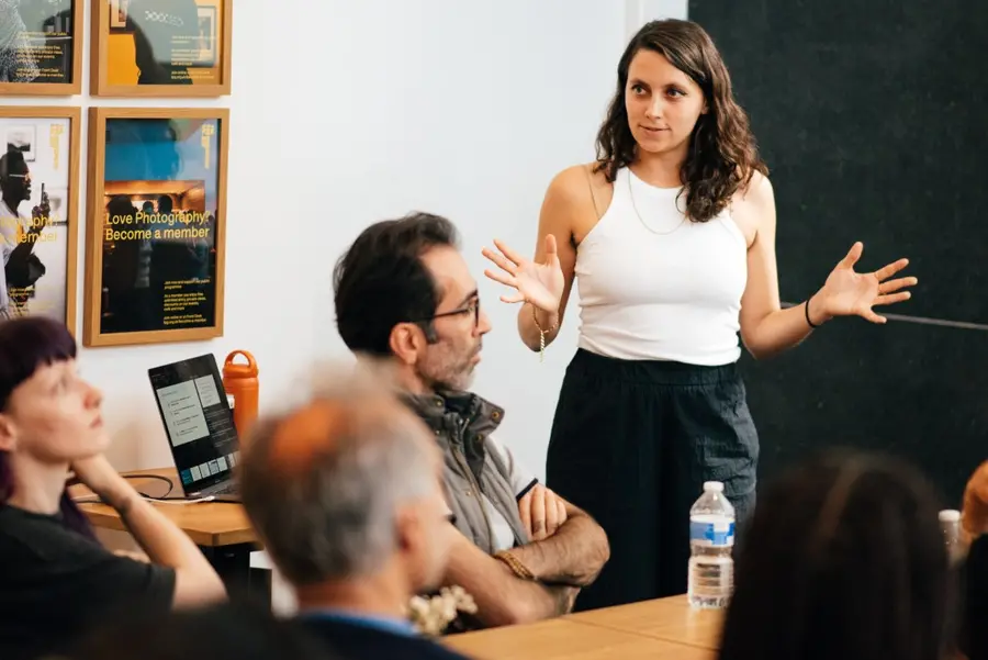 Colour photograph of a person standing at the front of the room, hands in an outward embrace, talking to people seated in front