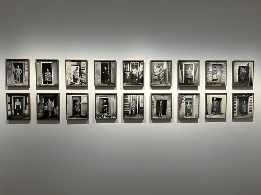 S series of framed black and white photographs hung in an exhibition space on a white wall. They all feature various solo women stood in the doorways to their homes.