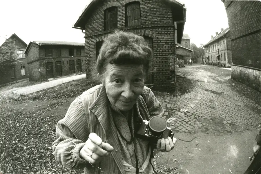 A black and white image of the Polish photographer, Zofia Rydet. She stares directly into the lens with a playful smirk on her face while she holds a camera in her left hand.